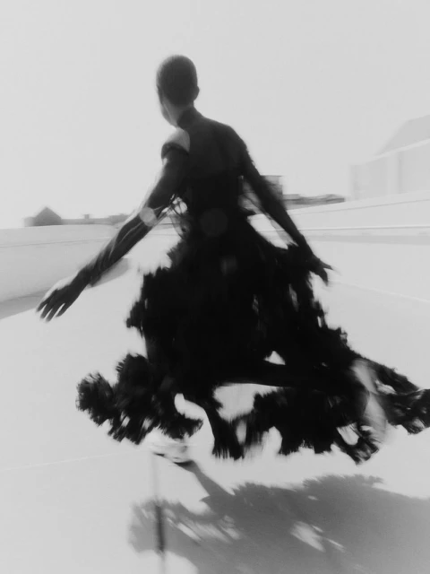 Monochrome image featuring a person in motion wearing a dark, flowing gown against a light neutral backdrop. The picture evokes a sense of motion.