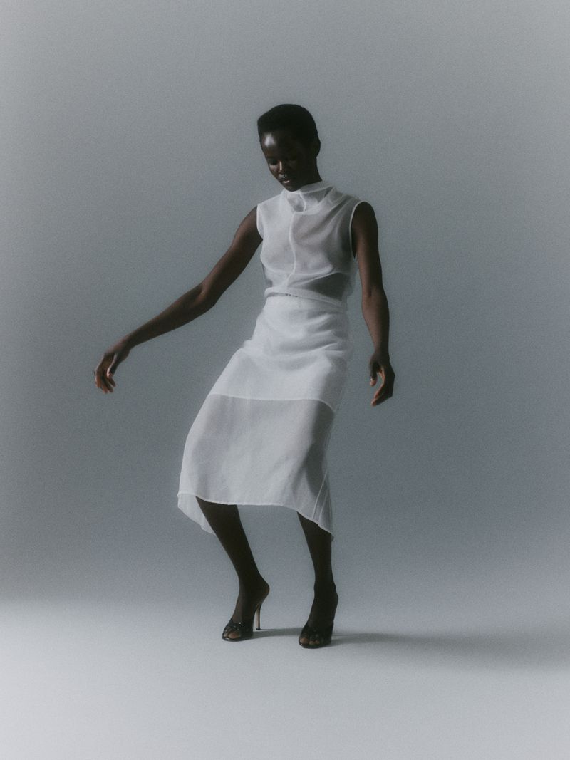 Fashion shot of a Black woman in a white see-through dress and heels posing in a photo studio with soft, diffused lighting.