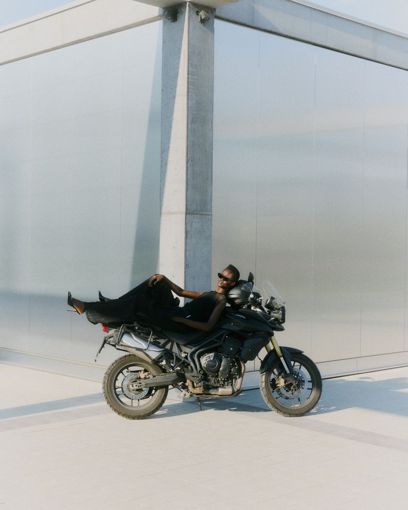 Woman in sunglasses and black dress lounging on a black motorcycle parked in front of modern building with concrete and metal exterior.