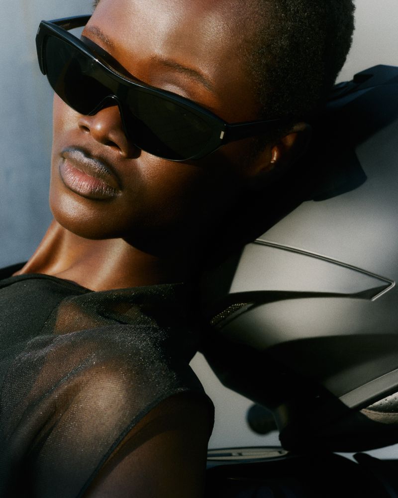 Close-up of a model wearing dark sunglasses and a black top, leaning against a motorcycle helmet in bright sunlight.
