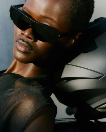 Close-up of a model wearing dark sunglasses and a black top, leaning against a motorcycle helmet in bright sunlight.