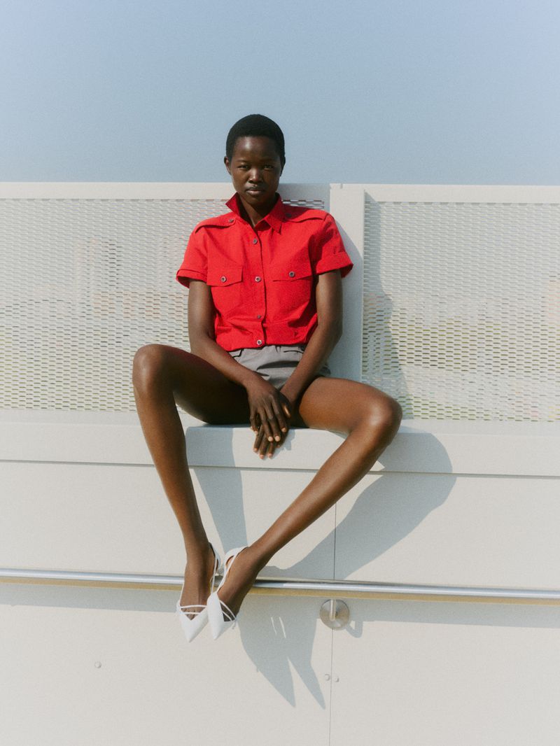 A striking dark-skinned model sits on a ledge against the sky in a red blouse, grey shorts, and white heels showcasing a fashion editorial shot.