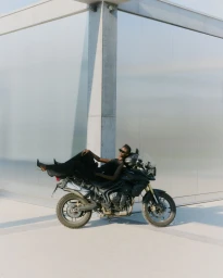 Model wearing black clothing reclining horizontally across a parked motorcycle before a concrete building exterior