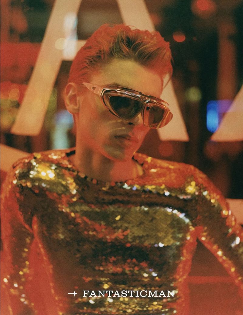 Adult male wearing futuristic sunglasses and a sparkling gold shirt poses indoors with red lighting and a modern, stylish aesthetic.