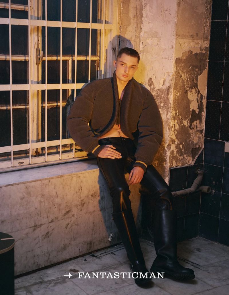 A male model poses seated against a window wearing a dark puffer jacket, leather pants, and boots in an indoor setting with neutral tones.