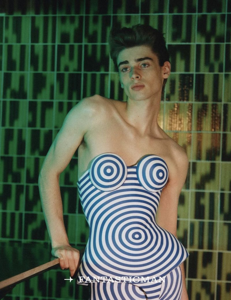 Striking studio portrait of a young man in a blue and white spiral-patterned outfit against a patterned green tile background, leaning on a rod.