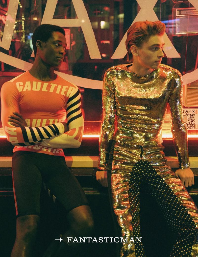 Two men pose indoors; one in gold sequin top and polka dot pants, the other in a 'Gaultier' top and athletic shorts, fashion shot.