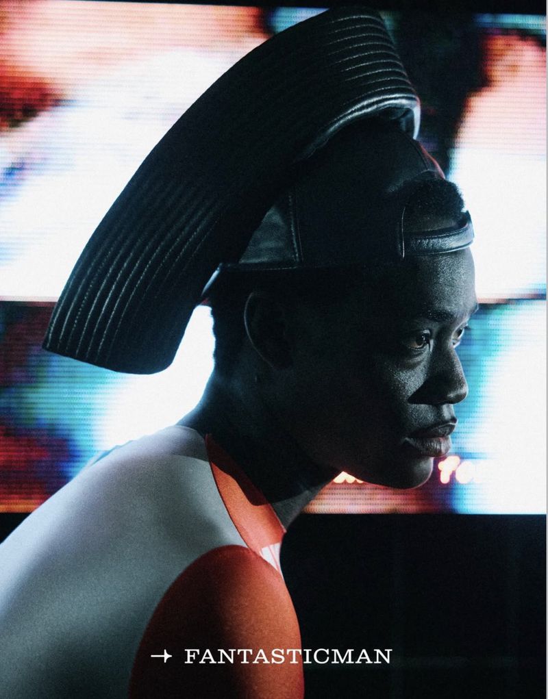 Profile view of a man wearing an architectural headpiece, styled in orange and white athletic wear in front of a colorful blurred backdrop.