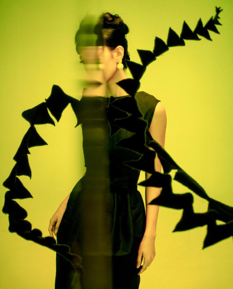 A stylish female fashion model poses in a green studio, wearing a dark vintage dress and large earrings, with a triangular pattern.