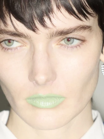 Close-up shot of a woman with light skin, dark hair, light eyes and green lipstick, wearing a white collared shirt and earring.