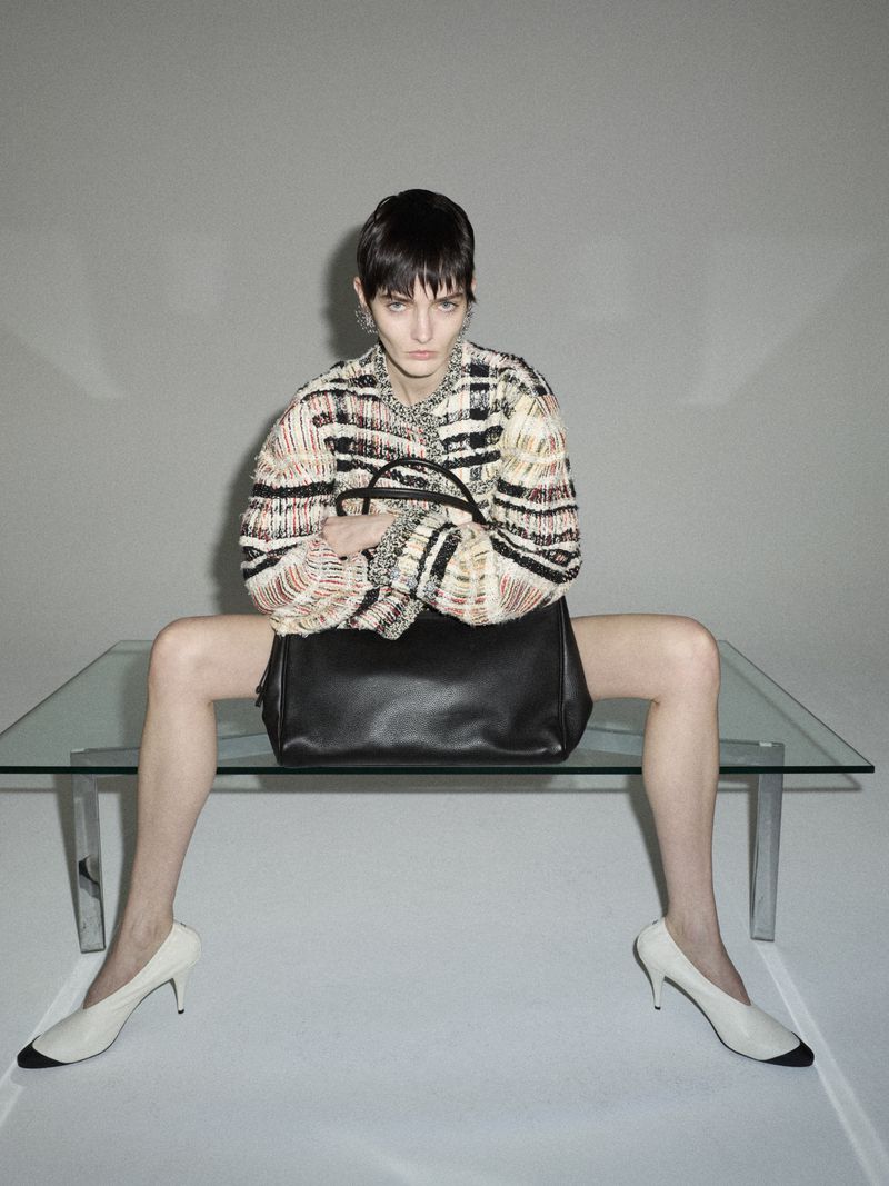 A stylish adult female model poses indoors, seated on a glass table with a black leather handbag, wearing a multi-colored woven jacket and cream heels.