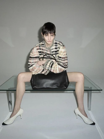 A stylish adult female model poses indoors, seated on a glass table with a black leather handbag, wearing a multi-colored woven jacket and cream heels.