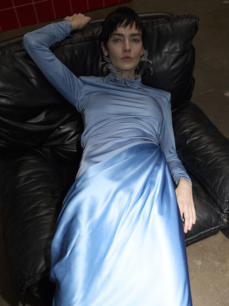 A woman lies on a black leather couch wearing a long-sleeved blue dress with a satin skirt, looking directly at the camera.