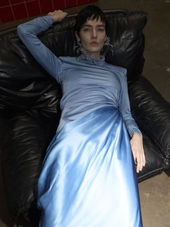 A woman lies on a black leather couch wearing a long-sleeved blue dress with a satin skirt, looking directly at the camera.