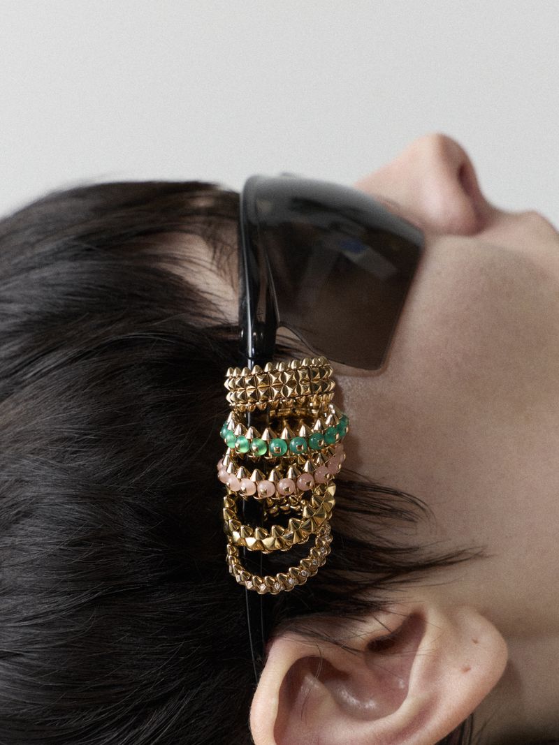 Close-up of a dark-haired human with multi-tiered gold, green, and pink jewelry adorning their hair near the ear, plus black sunglasses.