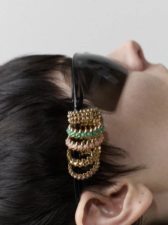 Close-up of a dark-haired human with multi-tiered gold, green, and pink jewelry adorning their hair near the ear, plus black sunglasses.
