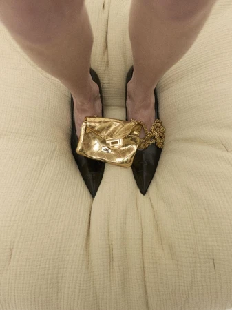Overhead shot of legs wearing black heels, a gold chain purse resting on the shoes, all against a textured cream fabric background.