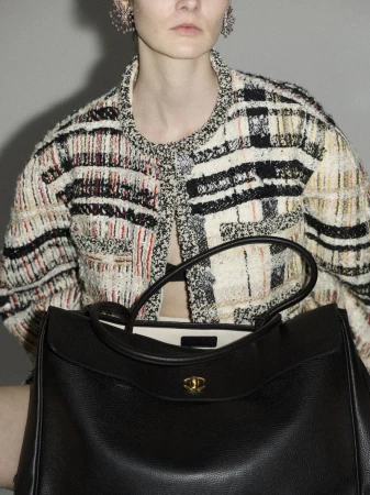 Woman poses with a large black leather tote bag, showcasing a patterned tweed jacket with sparkly earrings against a neutral backdrop.