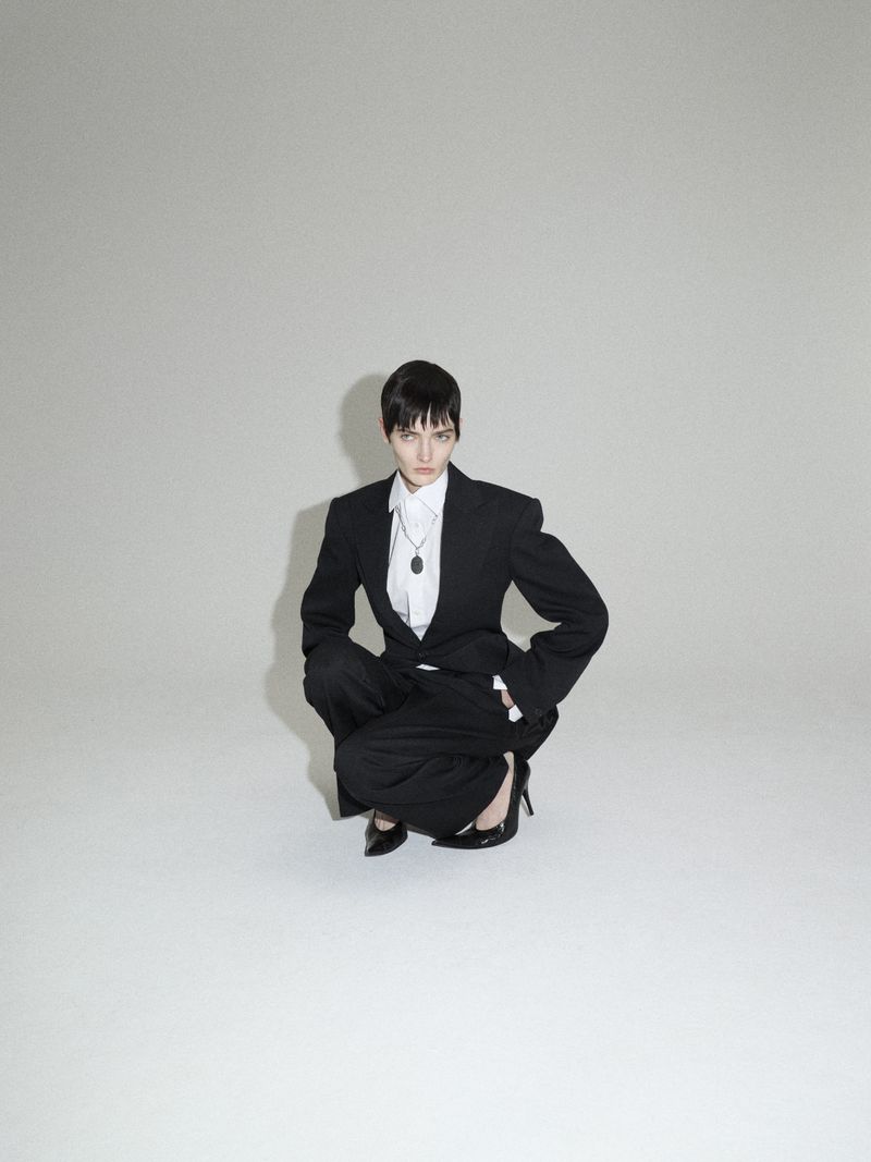 Woman in a black blazer and wide-leg pants crouches in a studio, wearing a white shirt, dark pendant necklace, and black high heels.
