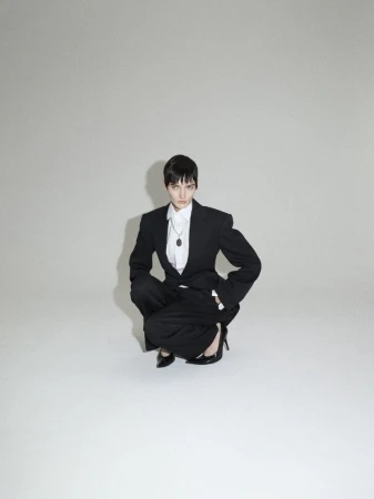 Woman in a black blazer and wide-leg pants crouches in a studio, wearing a white shirt, dark pendant necklace, and black high heels.