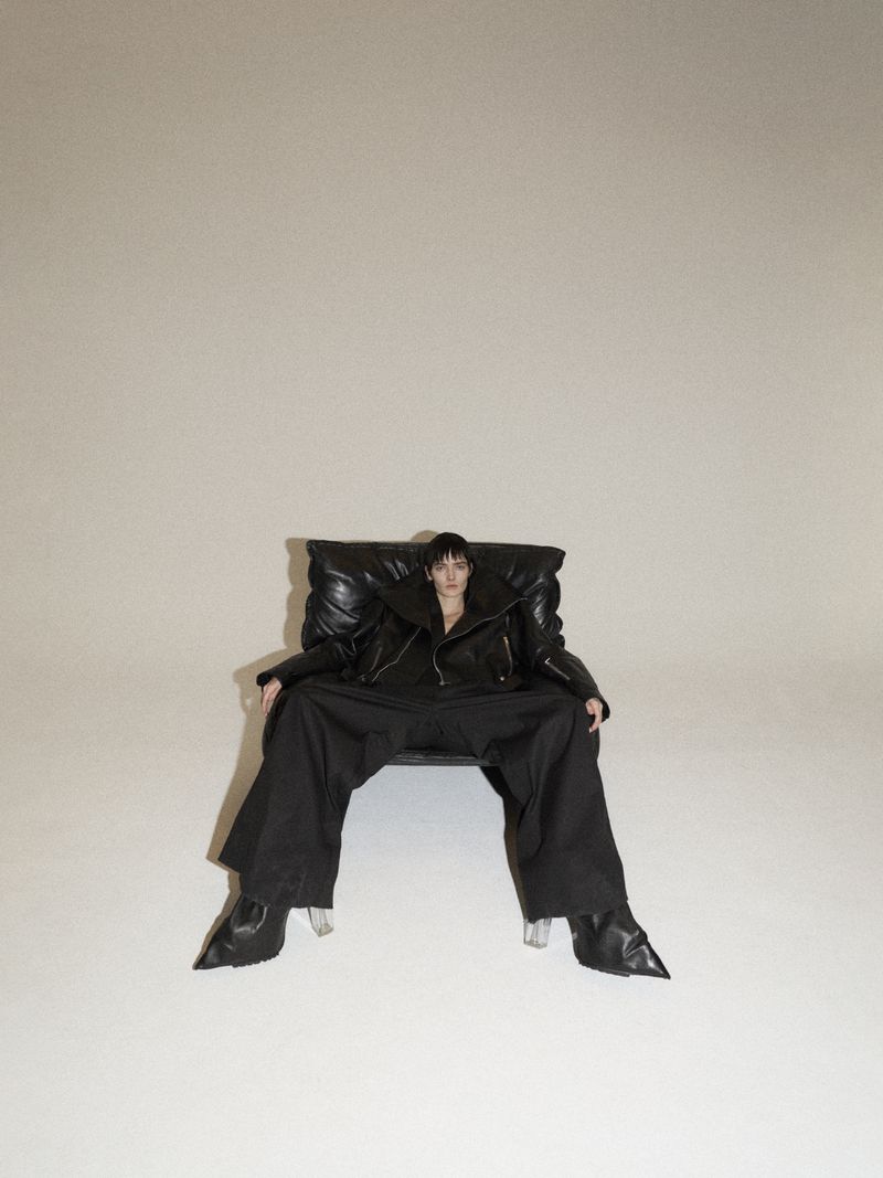 Woman in black leather jacket and wide-leg pants sits in a padded chair against a neutral backdrop in a fashion studio shot.