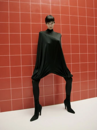 A solo female model with a short haircut in a full-body black fashion garment, standing indoors against a red-tiled wall on a white floor.