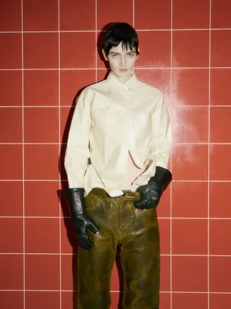 A person poses in front of a red tiled wall, wearing a yellow button-down shirt, distressed olive-green leather pants, and black leather gloves.