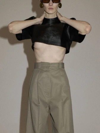 An adult female posing indoors, wearing a black leather crop top, tan wide-leg trousers, brown sunglasses and earrings against a plain background.
