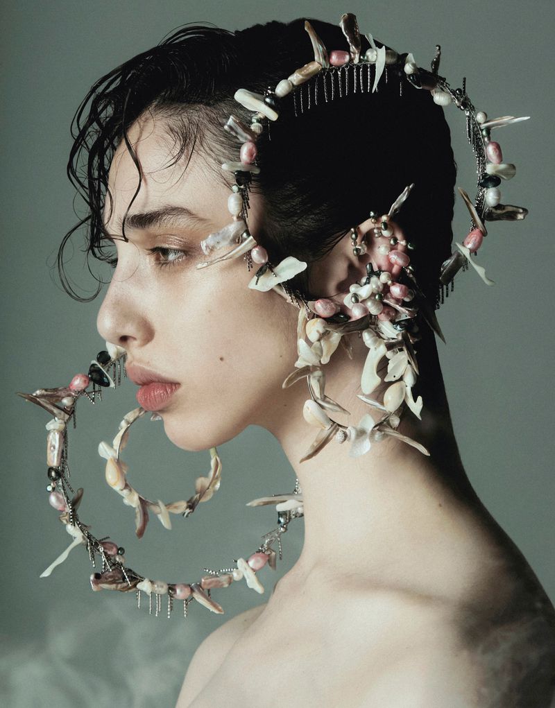Profile portrait of a young adult woman wearing elaborate jewelry made of shells and pearls in a studio setting with soft lighting.