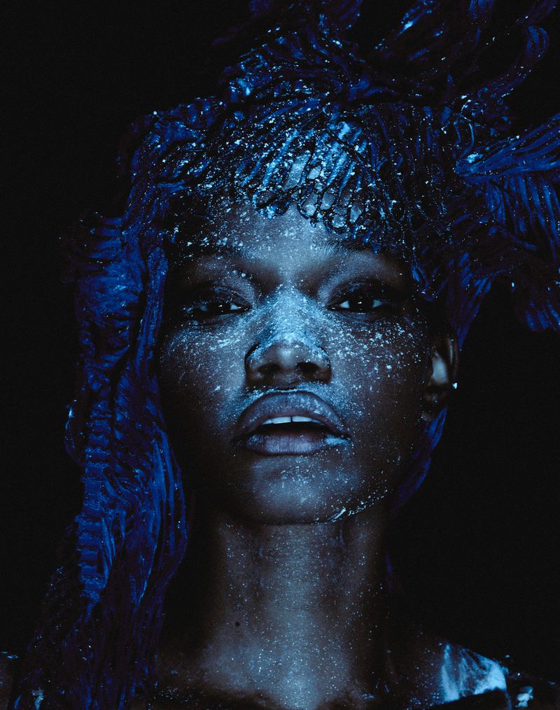 Close-up portrait of a Black woman with blue lighting and glitter on her face and decorative hair, set against a dark background.
