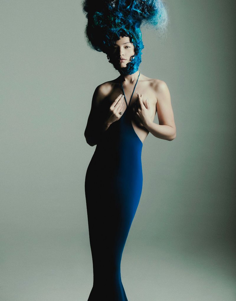 Woman in elegant blue dress with artistic blue hairpiece against a neutral background, posing with hands near chest, full shot.