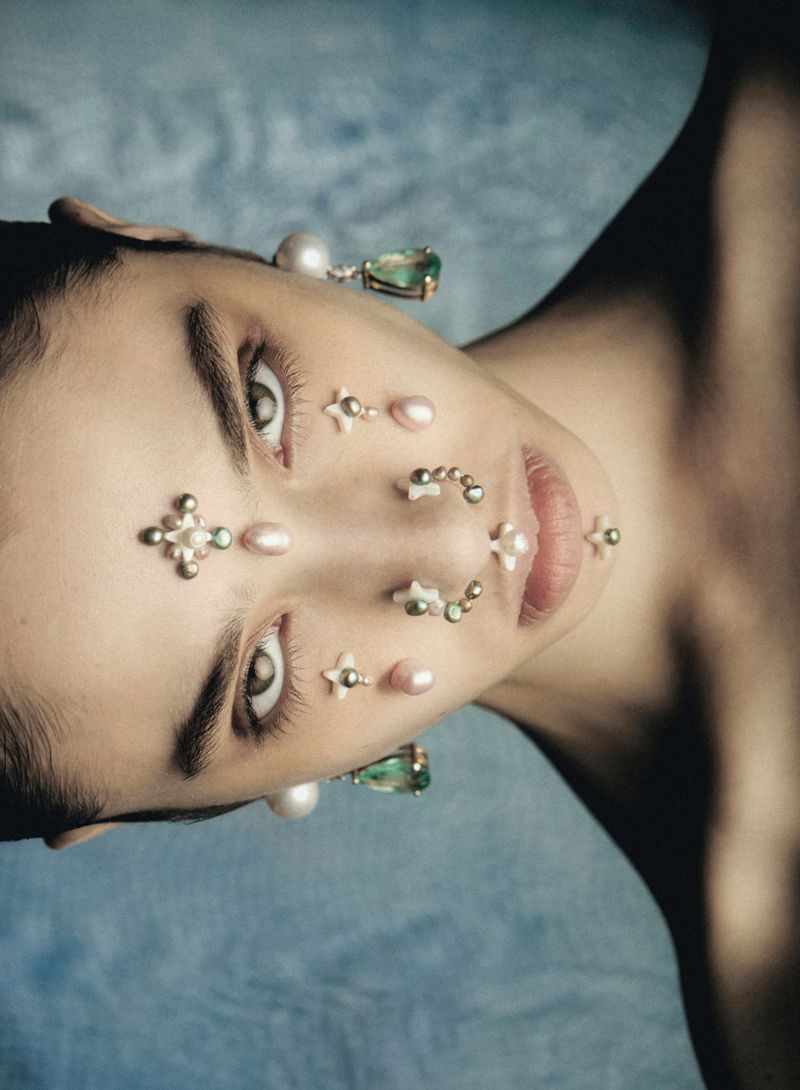 Close-up beauty shot of a light-skinned adult woman with pearls and star-shaped gems on her face against a soft blue backdrop.