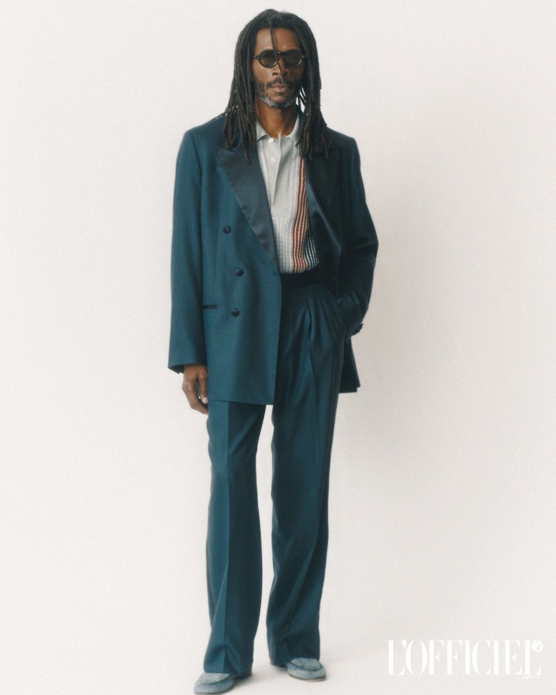 A male fashion model in a teal double-breasted suit with sunglasses, standing in a photo studio against a white backdrop, showcasing a modern style.
