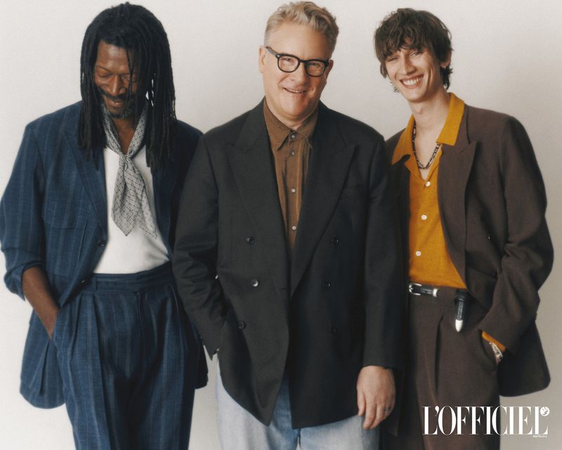 Group studio shot of three adult men in jackets and trousers, two smiling. Fashion editorial with studio lighting and neutral background.