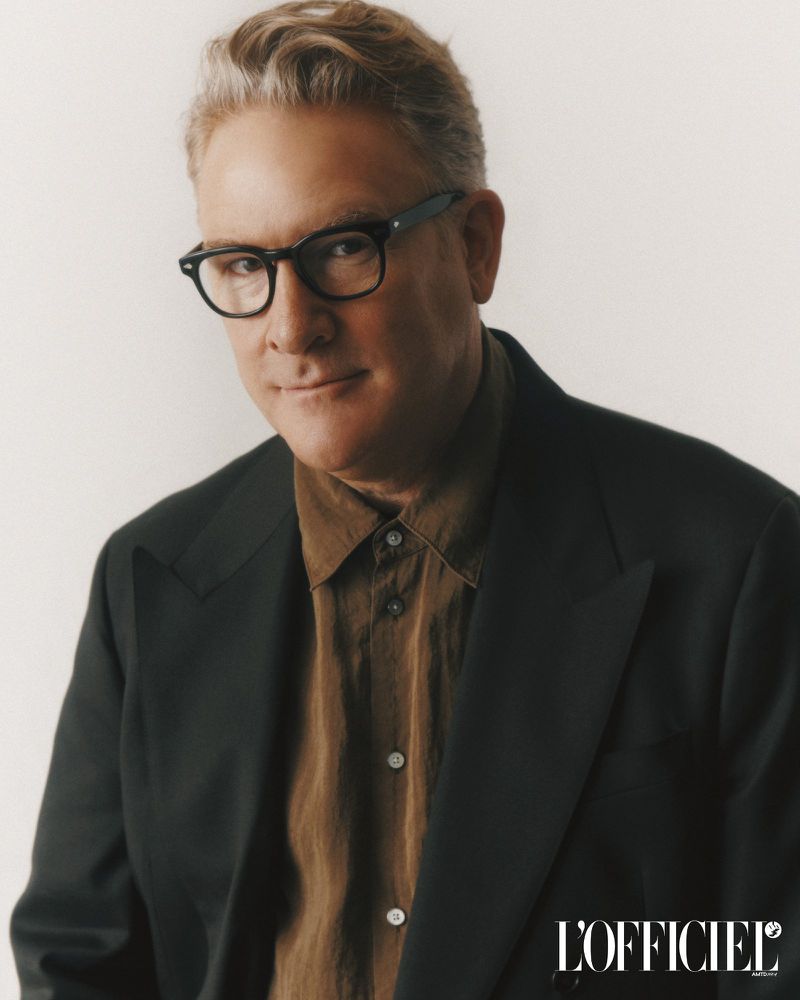 Portrait of an adult male with glasses wearing a brown shirt and black blazer in front of a white studio background.