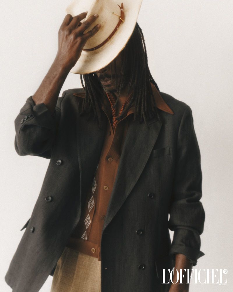 Adult male in stylish brown shirt and gray blazer, accessorized with a necklace and light colored cowboy hat, posing indoors.