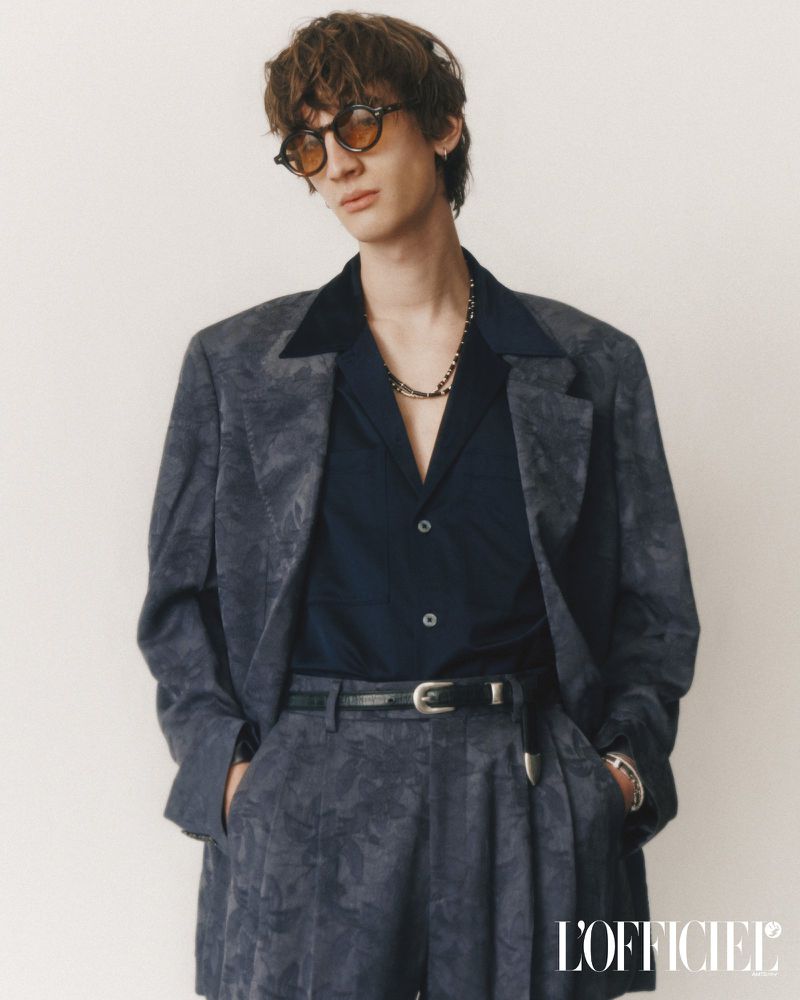 Fashion portrait of a young adult male with sunglasses wearing a patterned blazer, button-down shirt, and shorts against neutral backdrop.