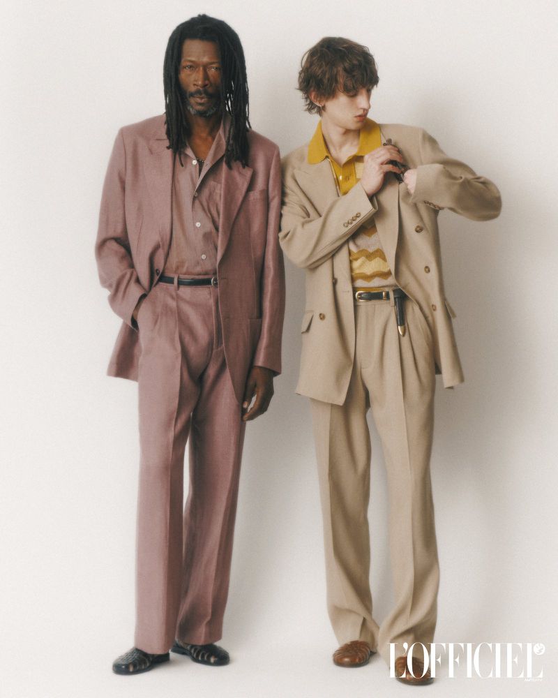 Two men stand indoors wearing fashionable blazers and trousers. The first man has dreadlocks, the second man has dark blonde hair.
