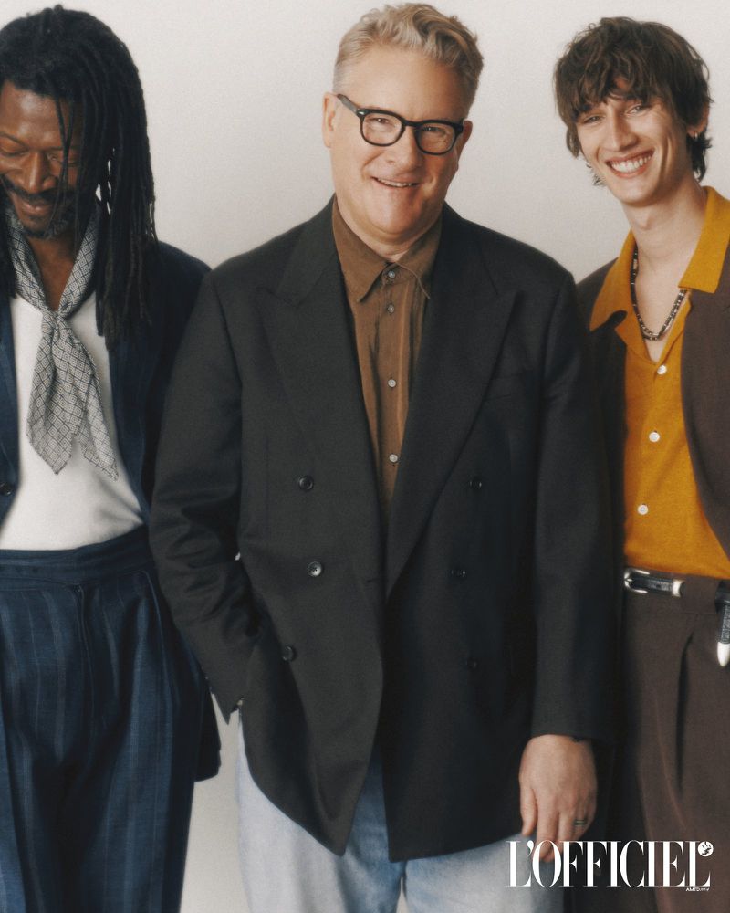 Group shot of three well-dressed men indoors. One is light-skinned wearing glasses and a dark blazer. The other two are in suits.