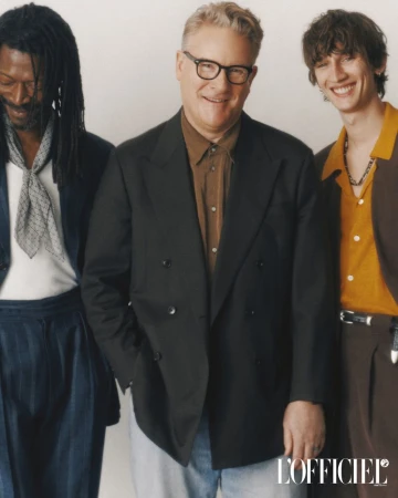 Group shot of three well-dressed men indoors. One is light-skinned wearing glasses and a dark blazer. The other two are in suits.
