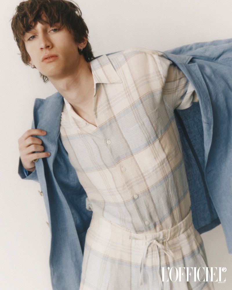 A young adult with dark hair poses in a plaid shirt and pants with a light blue jacket. The image is from L'Officiel magazine.
