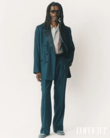A man with long dark dreadlocks wearing a teal suit jacket and matching trousers standing against a plain white background.