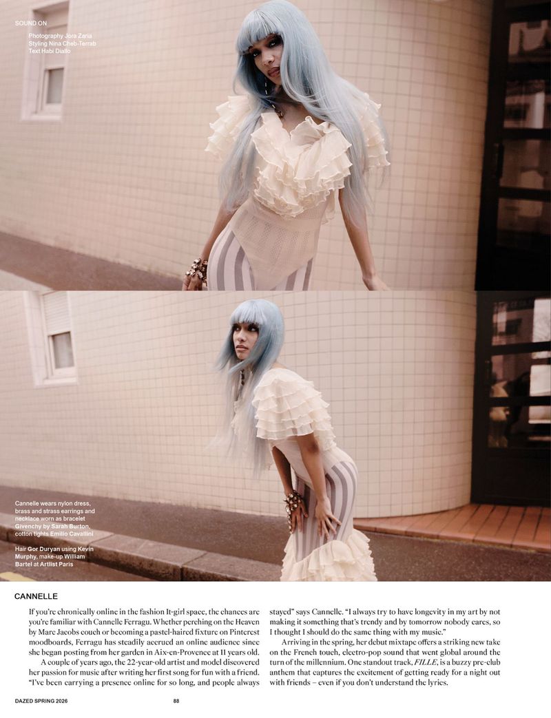 Artistic fashion portrait of a woman with light blue hair wearing a ruffled dress and bracelets, posing against a light-colored wall.