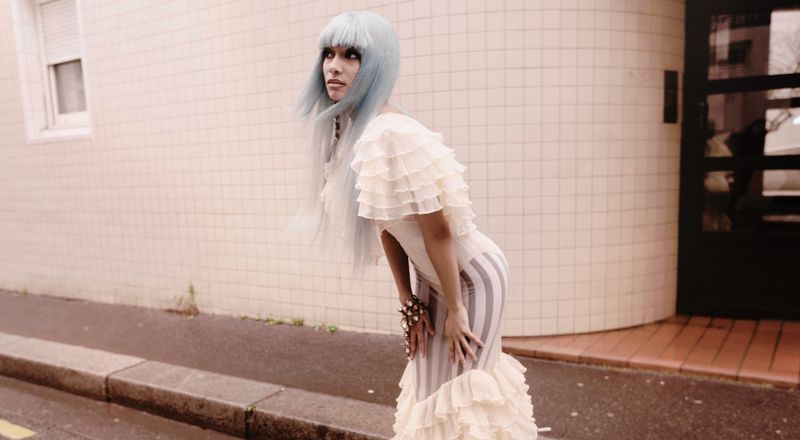 Woman in ruffled white dress and light blue wig poses on city street with neutral background near tiled wall and doorway.