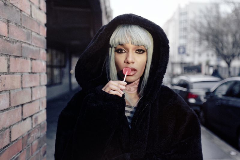 Stylish woman in a fur hood eating a lollipop in an urban setting, near a brick wall, with cars and buildings in the background.