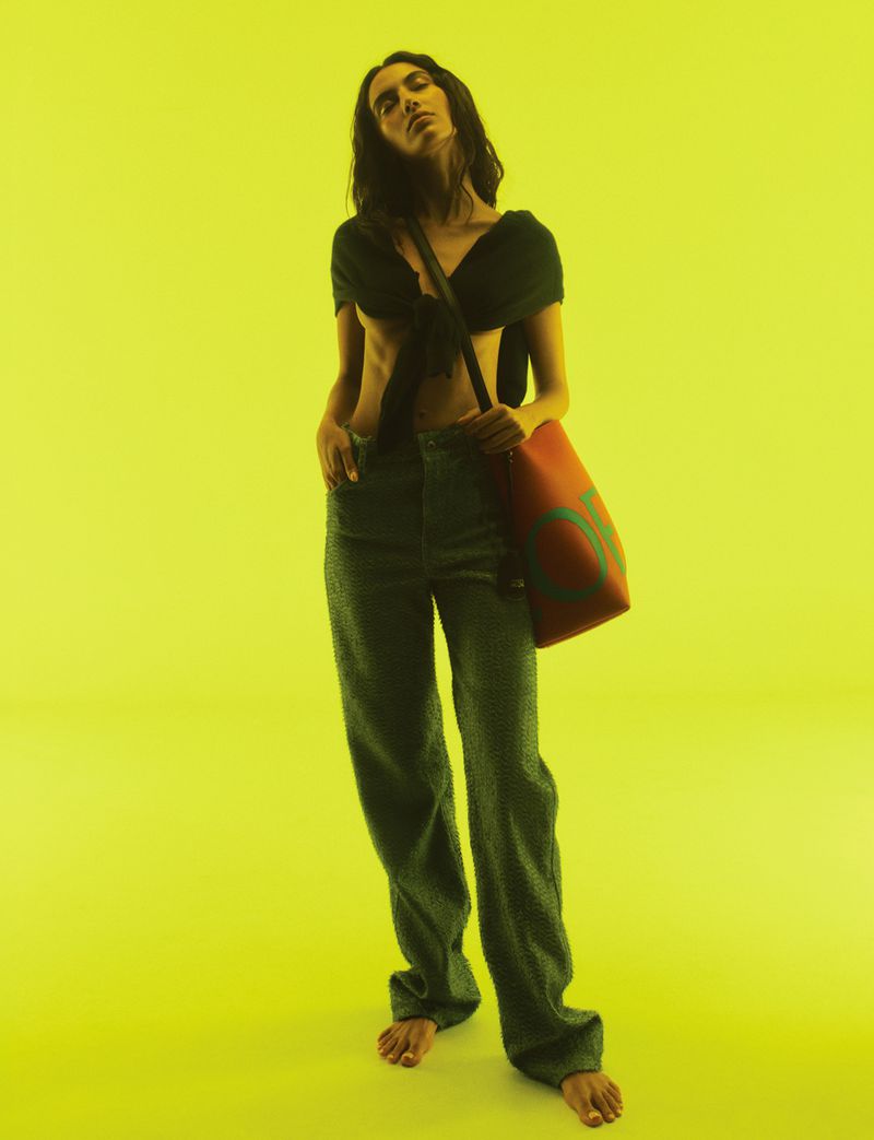 A barefoot adult woman wearing a black crop top and green jeans stands in a studio with a yellow backdrop and a red tote bag.