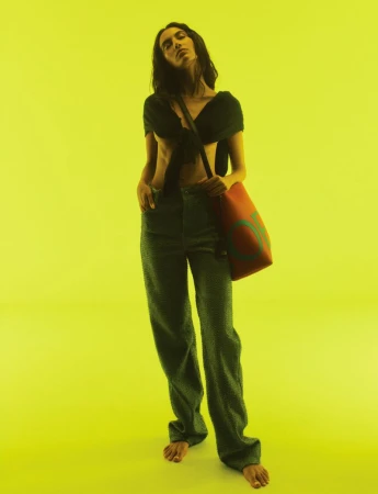 A barefoot adult woman wearing a black crop top and green jeans stands in a studio with a yellow backdrop and a red tote bag.