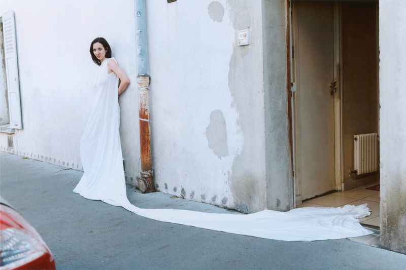 Woman in a long white dress standing against a pale street wall for SHADOWPLAY Magazine