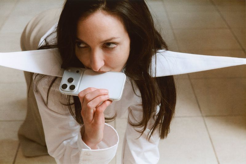 Woman in a white shirt poses on the floor, holding a smartphone to her mouth with three prominent camera lenses visible.