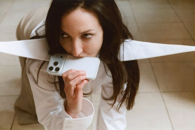 Woman in a white shirt poses on the floor, holding a smartphone to her mouth with three prominent camera lenses visible.
